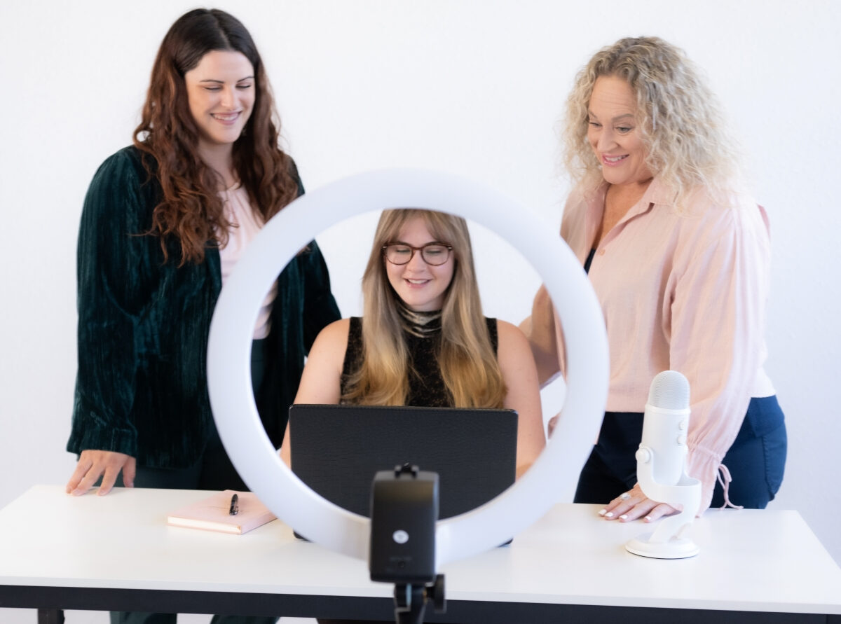 Three women collaboratively working around a laptop with a ring light and microphone, symbolizing the investment of hiring a virtual assistant.