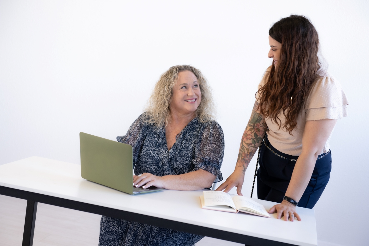 Two women smiling and working together at a desk with a laptop and notebook, exemplifying an organized and supportive environment.