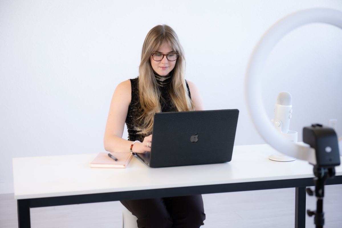 Focused woman with long blonde hair and glasses, working on a laptop with a microphone and ring light, setting herself up for success this fall.