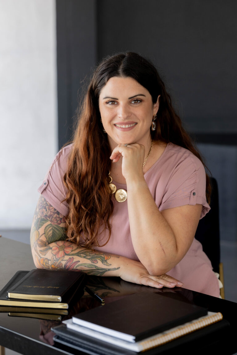 Smiling business consultant with long dark hair, wearing a pink top, sitting at a desk with notebooks, providing guidance on scaling your business.