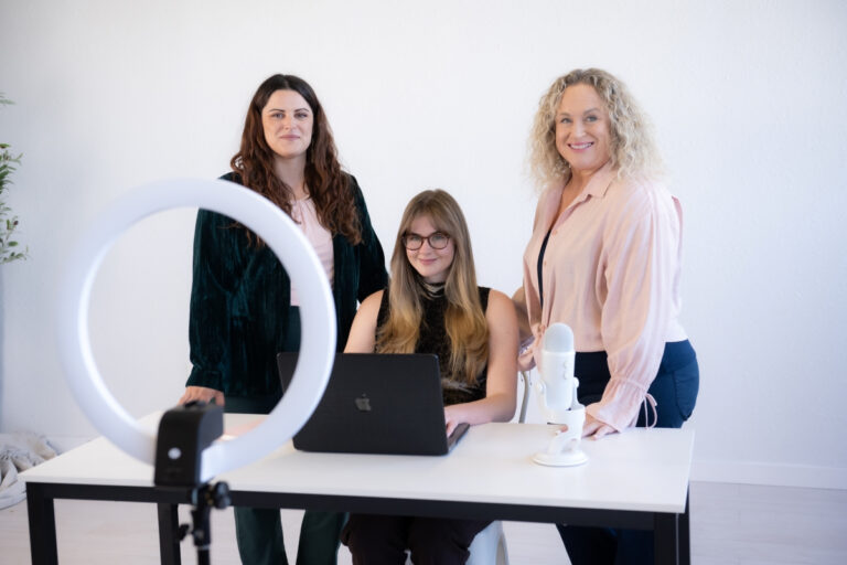 Three women collaborating in an office space, featuring a laptop, microphone, and ring light, highlighting a small business client success story.