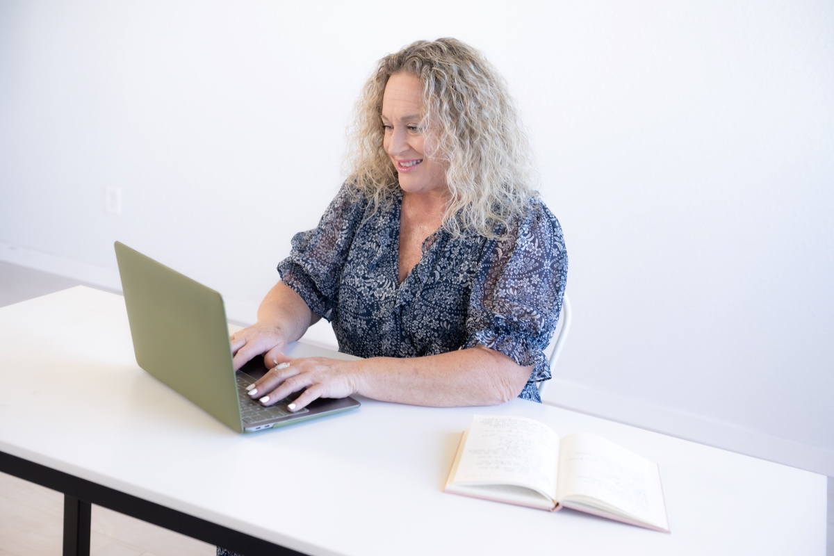 Smiling woman with curly hair typing on a green laptop at a desk with an open journal, representing productivity and VA tips for success.