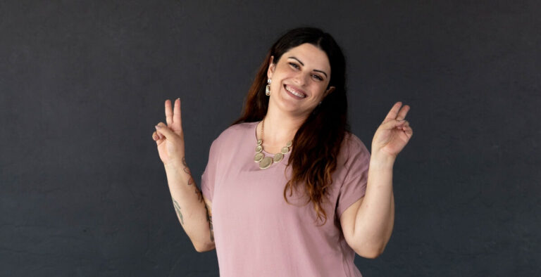 A smiling woman in a pink blouse and white pants poses with peace signs in front of a dark gray wall, featured on Canvas Rebel.