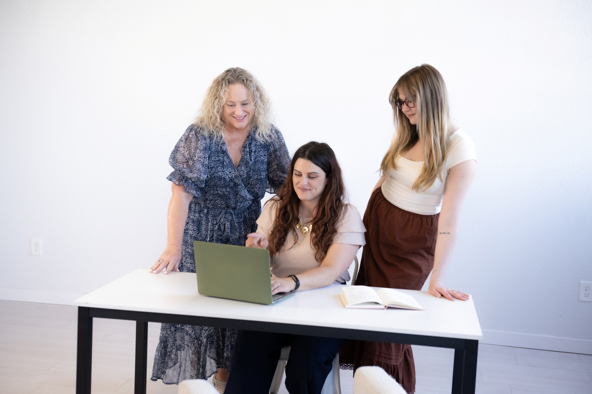 Three professional women collaborating at a desk with a laptop and notebook, discussing time management tips for improving business efficiency.