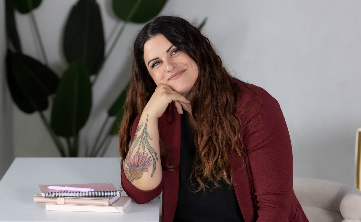 Smiling woman with long wavy hair, wearing a maroon blazer, sitting at a desk with notebooks—representing a professional virtual assistant ready to help scale your business.
