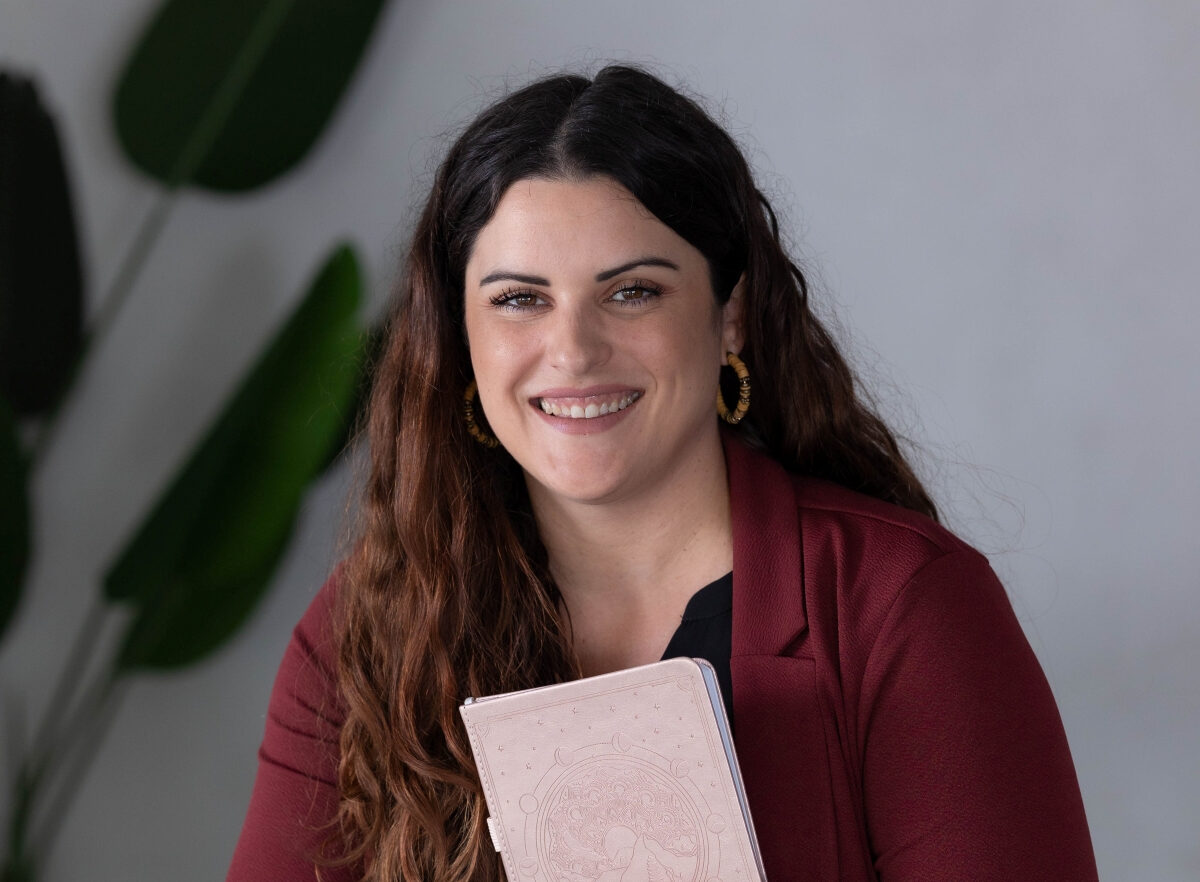 Speaker on Queens Lead podcast smiling and holding a pink journal, seated in a modern office space with greenery in the background.